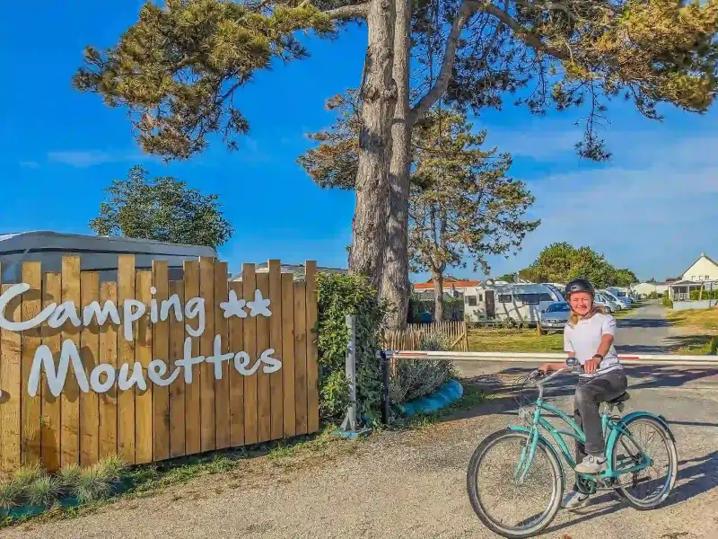 Vacancière à vélo devant l’entrée du camping Les Mouettes à Agon-Coutainville dans la Manche.